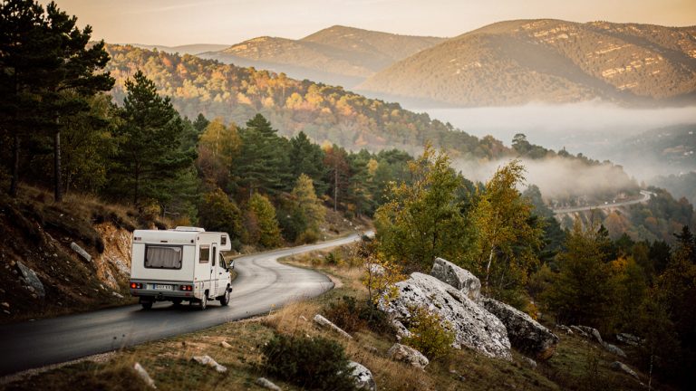 Un camping-car discret parcourt une petite route sinueuse des Cévennes au lever du soleil, entre forêt, brume légère et reliefs doux.